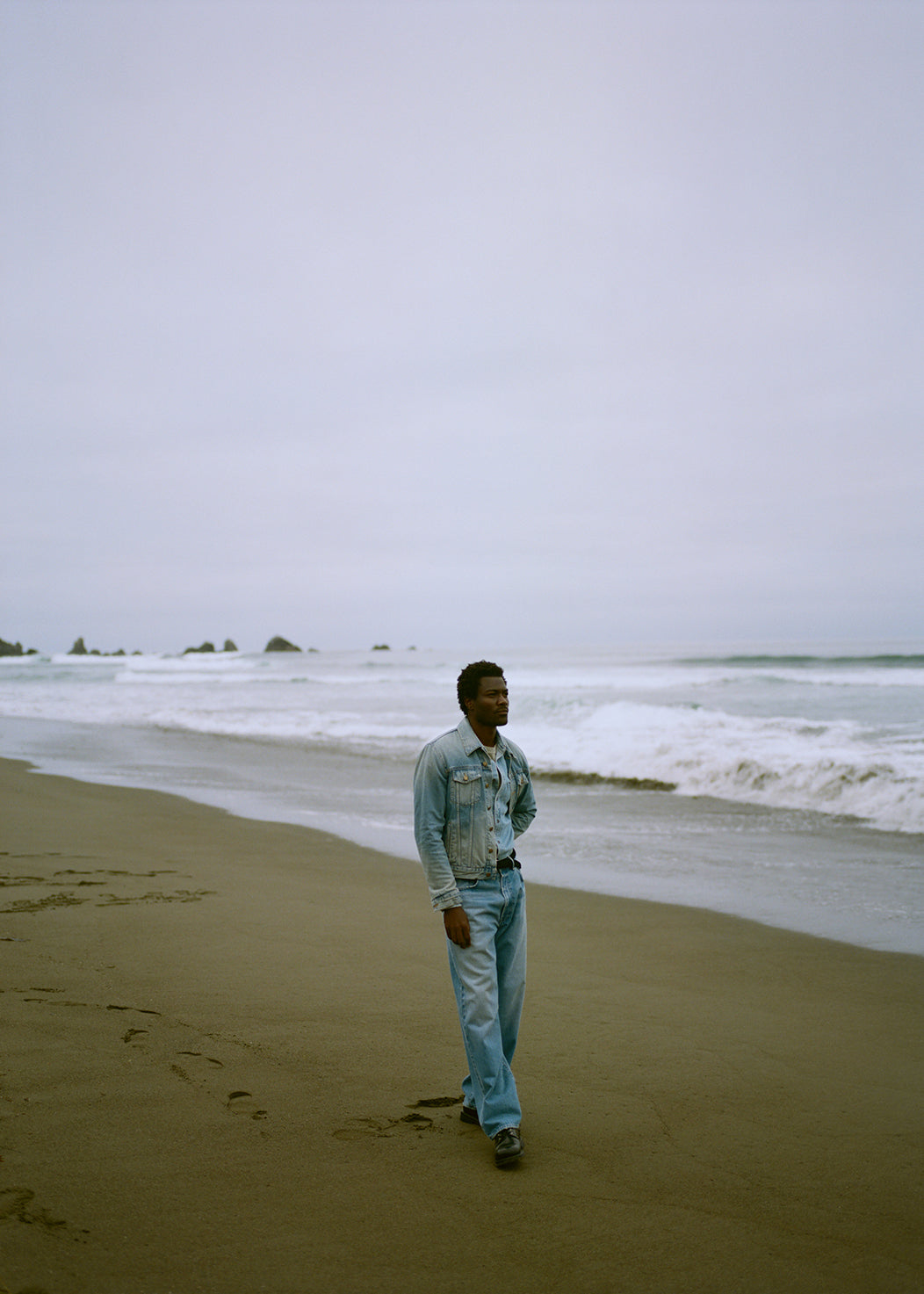 A man wearing Ninety One Baggy in Maldon, Brando Denim Jacket in Vintage Indigo, Long Sleeve Cairo Shirt in Charmer, and Everyday Short Sleeve Tee in Burnished Quartz on the beach