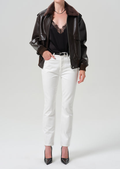 A model wearing a dark brown faux-leather jacket with a brown faux-shearling collar, styled with a black lace-trimmed top, white straight-leg jeans, and black pointed-toe heels, against a grey background.