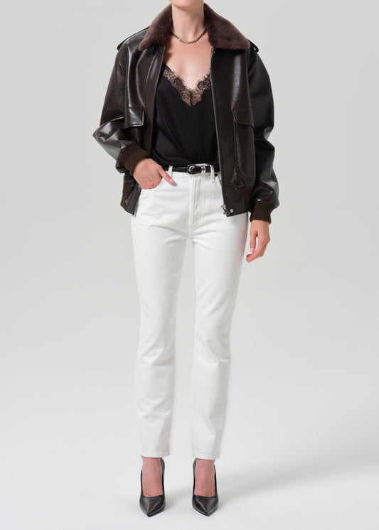 A model wearing a dark brown faux-leather jacket with a brown faux-shearling collar, styled with a black lace-trimmed top, white straight-leg jeans, and black pointed-toe heels, against a grey background.