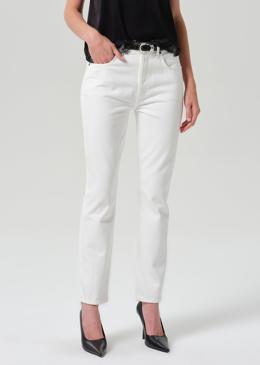 White slim-fit jeans worn by a model, styled with a black belt, black top, and black pointed-toe heels against a grey background.