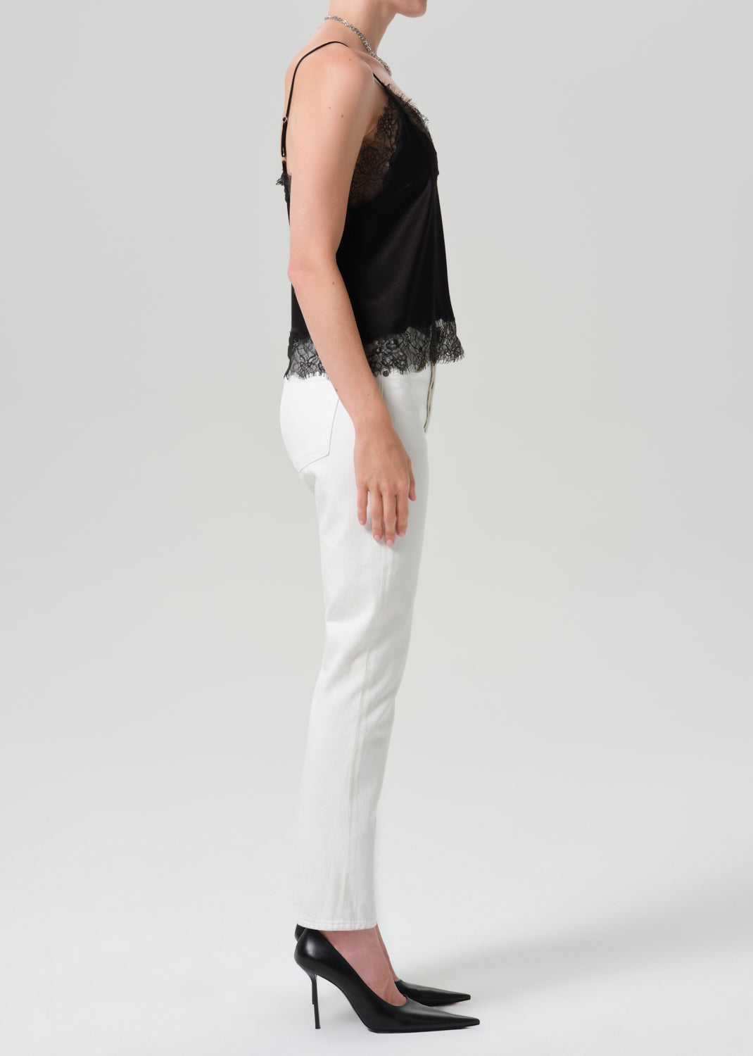A model shown from the side wearing a black camisole top with sheer lace panels and a lace hem, paired with white pants and black heels, against a grey background.
