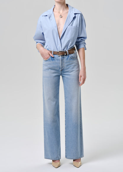 A model wearing a light blue button-down shirt, light blue wide-leg denim jeans, a brown leather belt, and beige pointed-toe heels, photographed against a grey background.