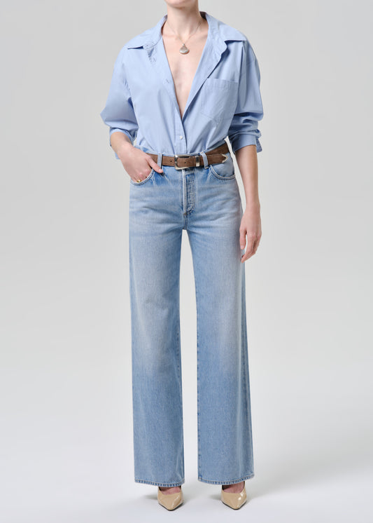 A model wearing a light blue button-down shirt, light blue wide-leg denim jeans, a brown leather belt, and beige pointed-toe heels, photographed against a grey background.