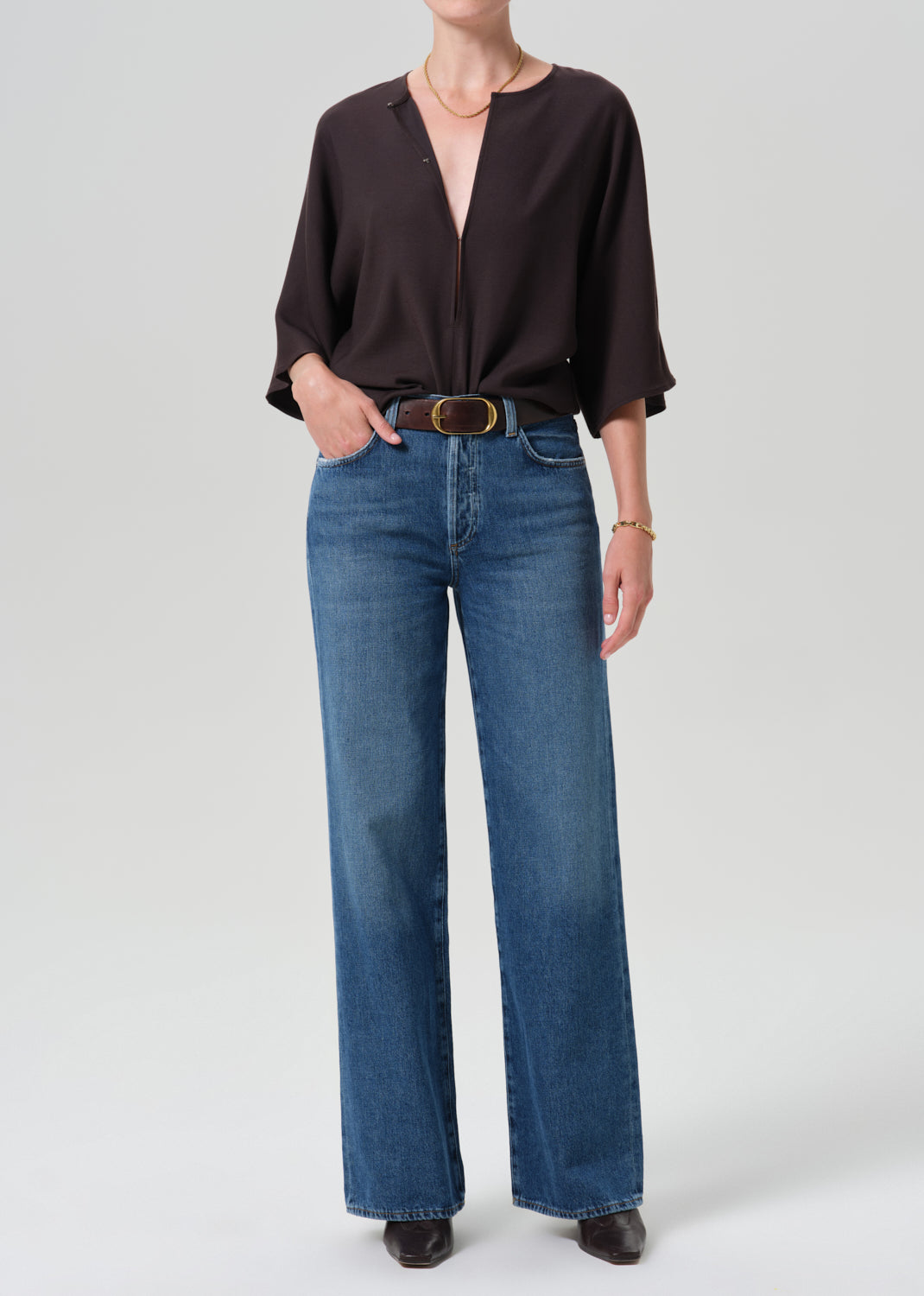 A model wearing a dark brown long-sleeve blouse tucked into high-waisted, wide-leg blue jeans with a gold-tone belt, standing against a grey background.