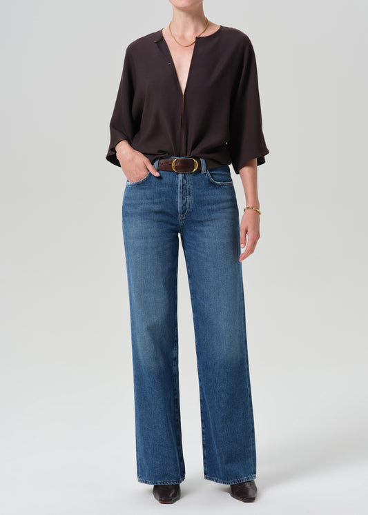A model wearing a dark brown long-sleeve blouse tucked into high-waisted, wide-leg blue jeans with a gold-tone belt, standing against a grey background.