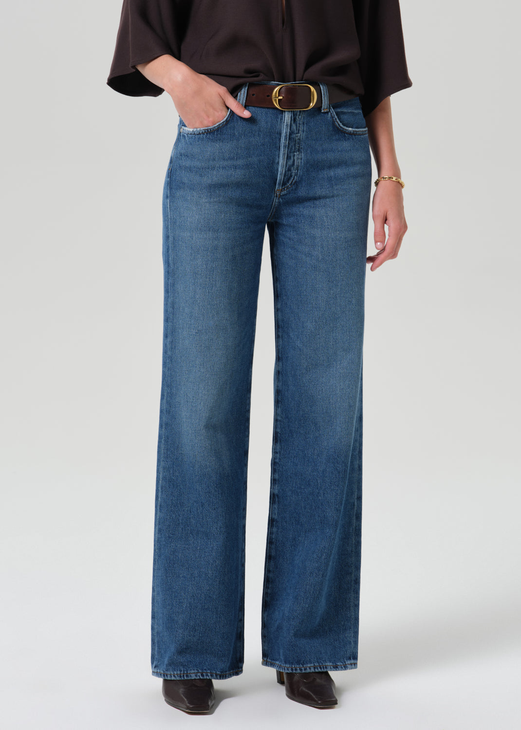 A model wearing a pair of wide-leg blue denim jeans with a dark brown shirt and a gold-buckled belt, photographed against a grey background.