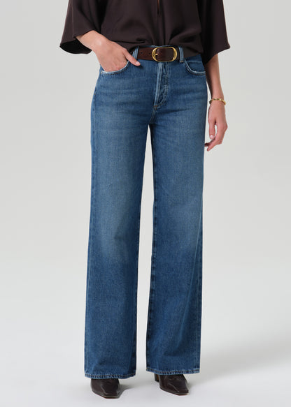 A model wearing a pair of wide-leg blue denim jeans with a dark brown shirt and a gold-buckled belt, photographed against a grey background.