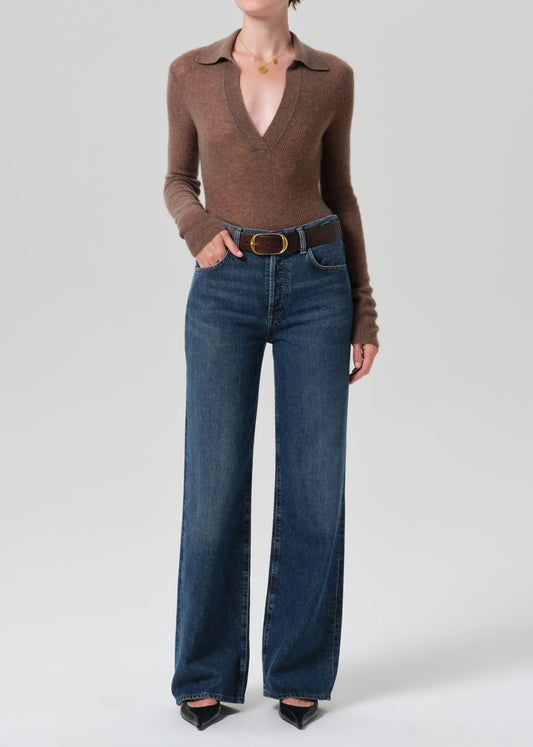 A model wearing a brown long-sleeve knit polo-neck top and high-waisted, wide-leg blue jeans with a brown belt, standing against a grey background.
