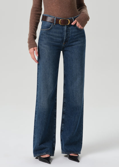 A model wearing a pair of dark blue wide-leg jeans with a brown belt, styled with a brown knit top and black pointed-toe heels against a grey background.