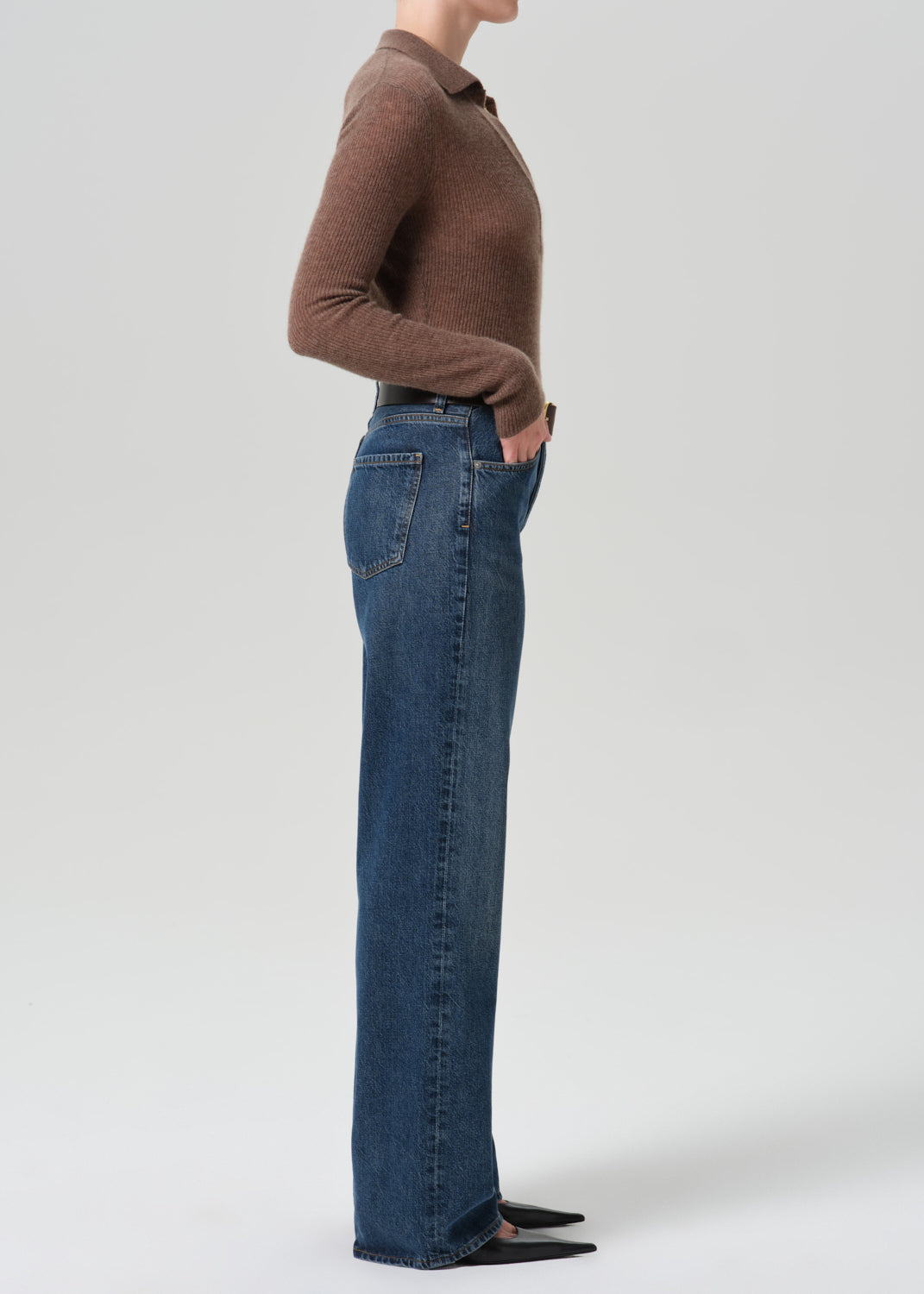 A side-profile studio shot of a model wearing a brown long-sleeve knit polo shirt and high-waisted, wide-leg blue denim jeans against a grey background.
