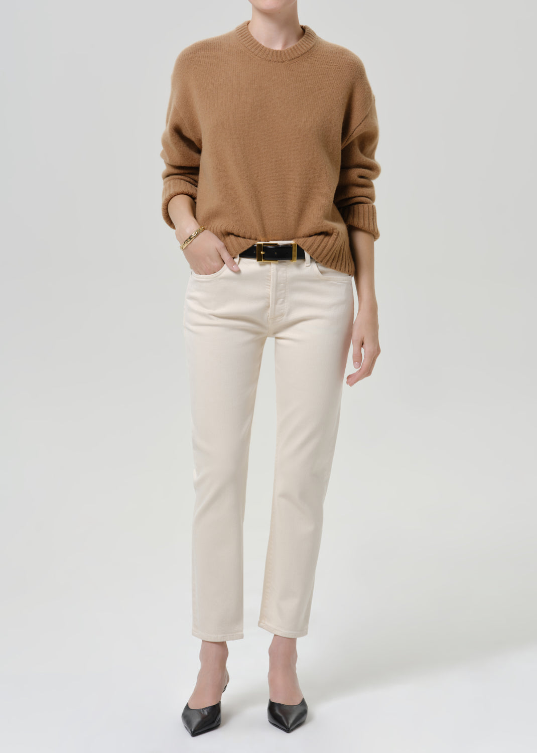 Person wearing cream straight leg cropped jeans with a camel brown crewneck sweater, black belt with gold buckle, and black pointed-toe heels, front full body view on a grey background.