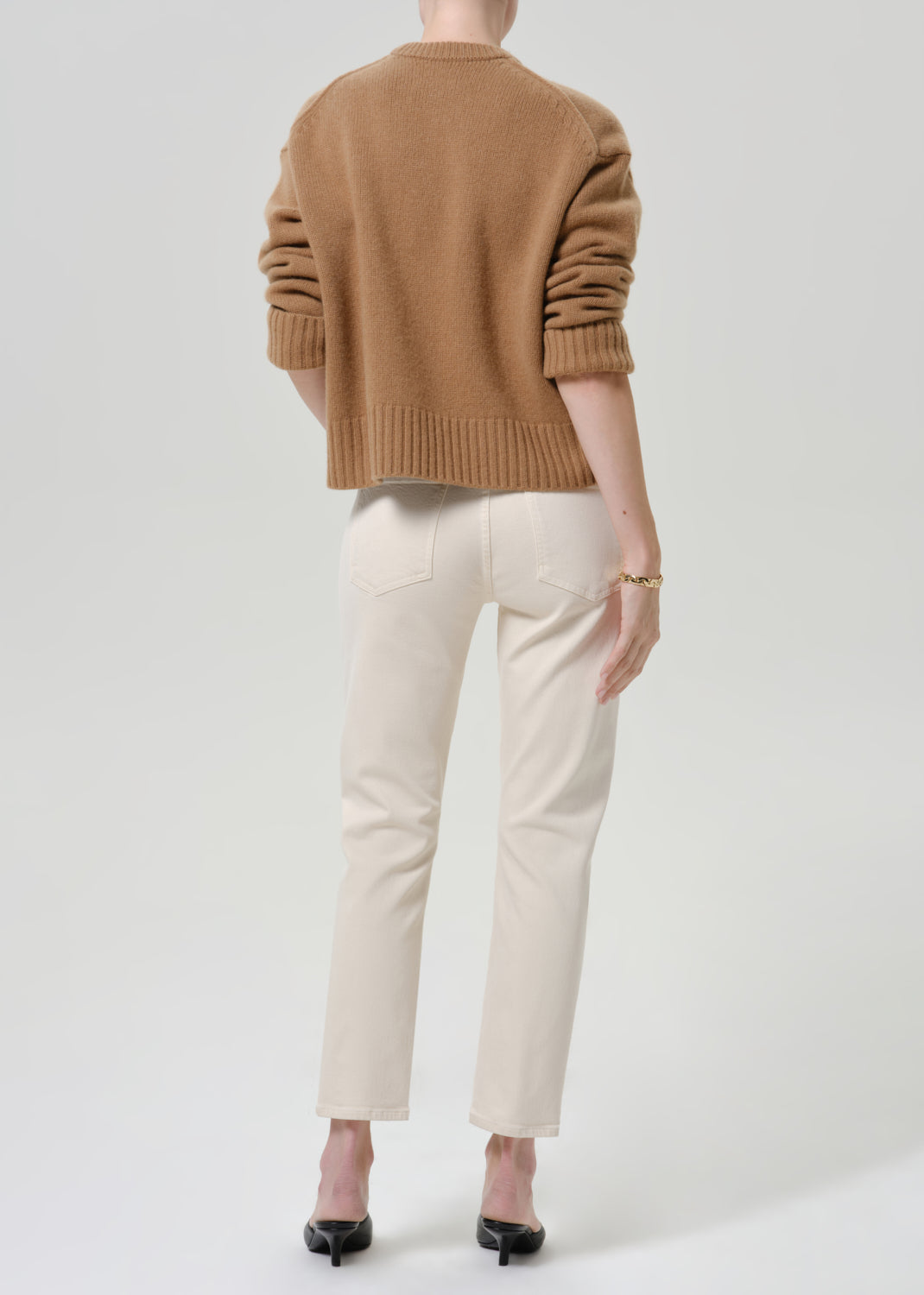 Person wearing cream straight leg cropped jeans with a camel brown crewneck sweater and black heels, back full body view on a grey background.