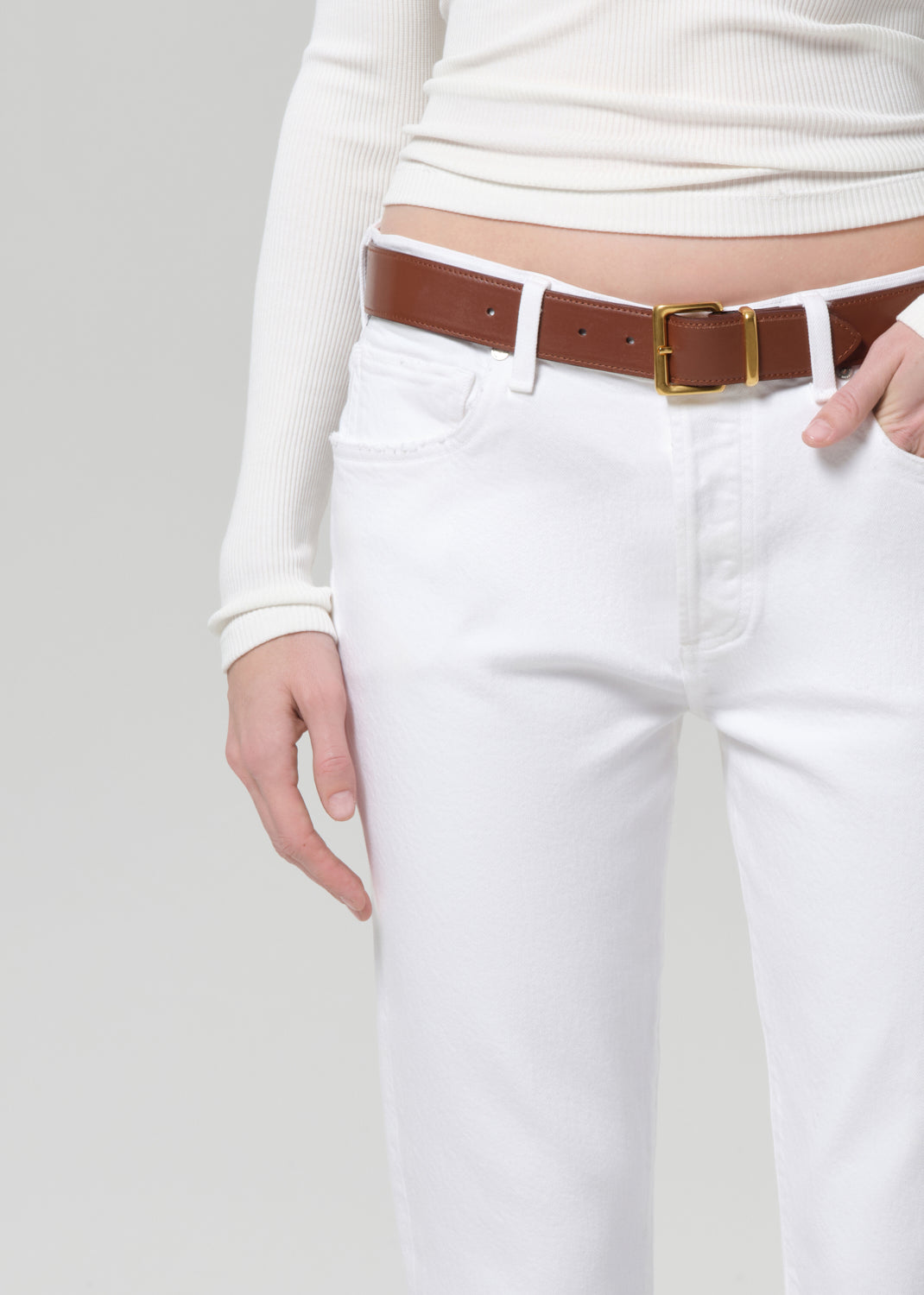 Person wearing a white long-sleeve top and white jeans with a brown belt on a light gray background