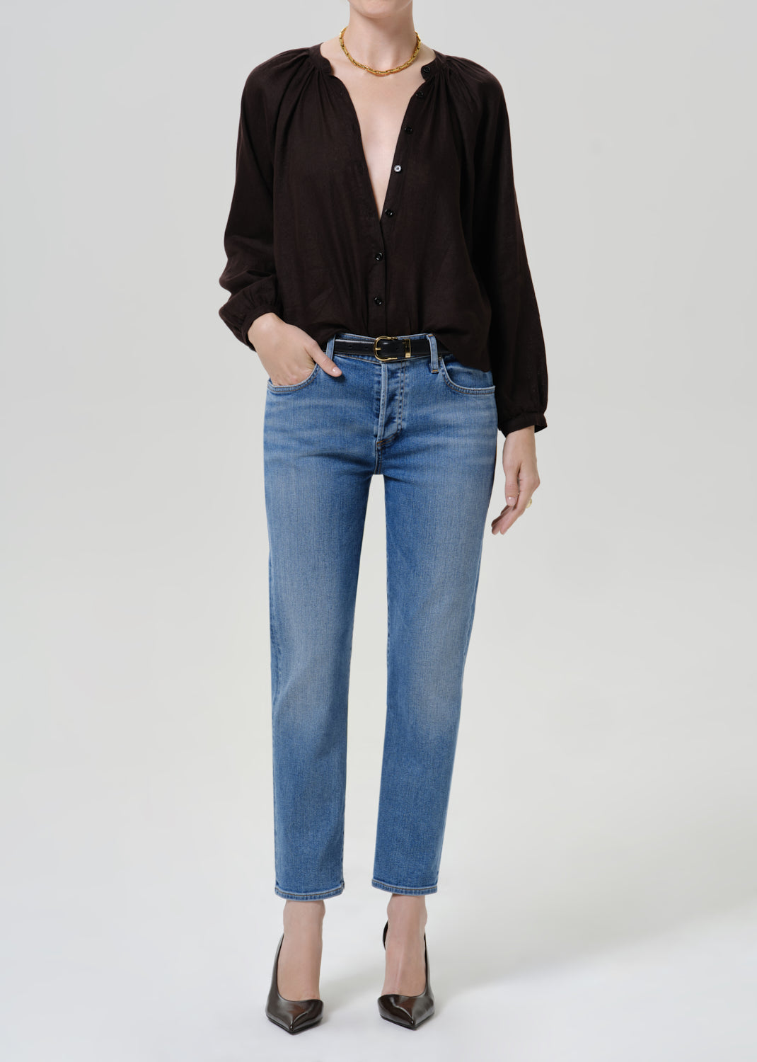Person wearing medium blue straight leg cropped jeans with a black long sleeve blouse, black belt with gold buckle, and black pointed-toe heels, front full body view on a grey background.
