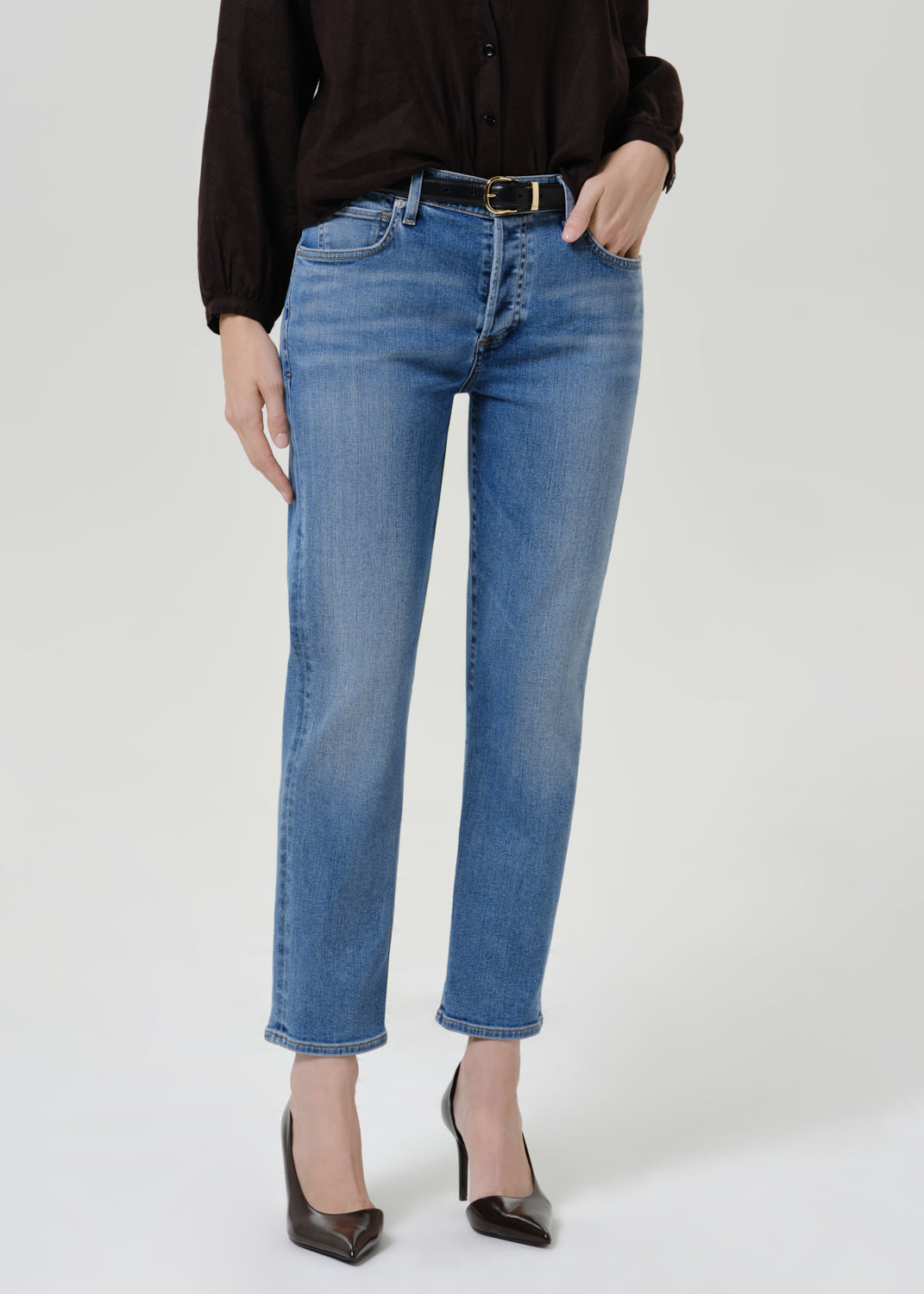 Person wearing medium blue straight leg cropped jeans with a black long sleeve blouse and black heels, front cropped view on a grey background.