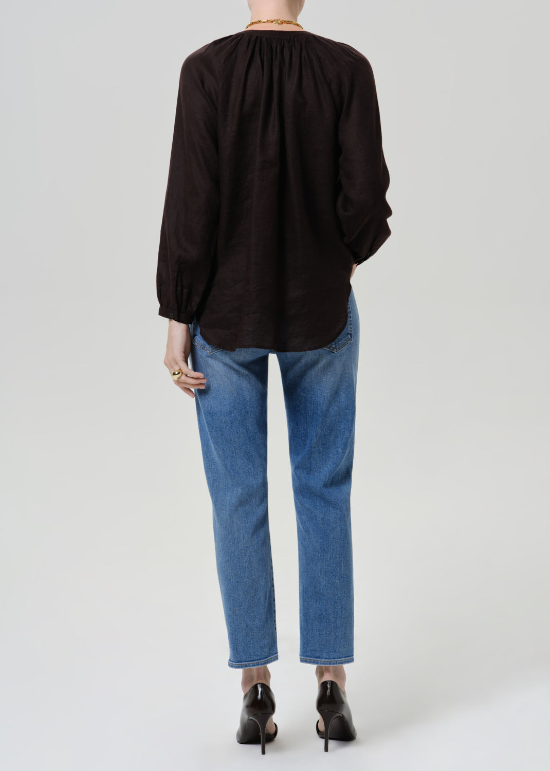 Person wearing medium blue straight leg cropped jeans with a black long sleeve blouse and black heels, back full body view on a grey background.