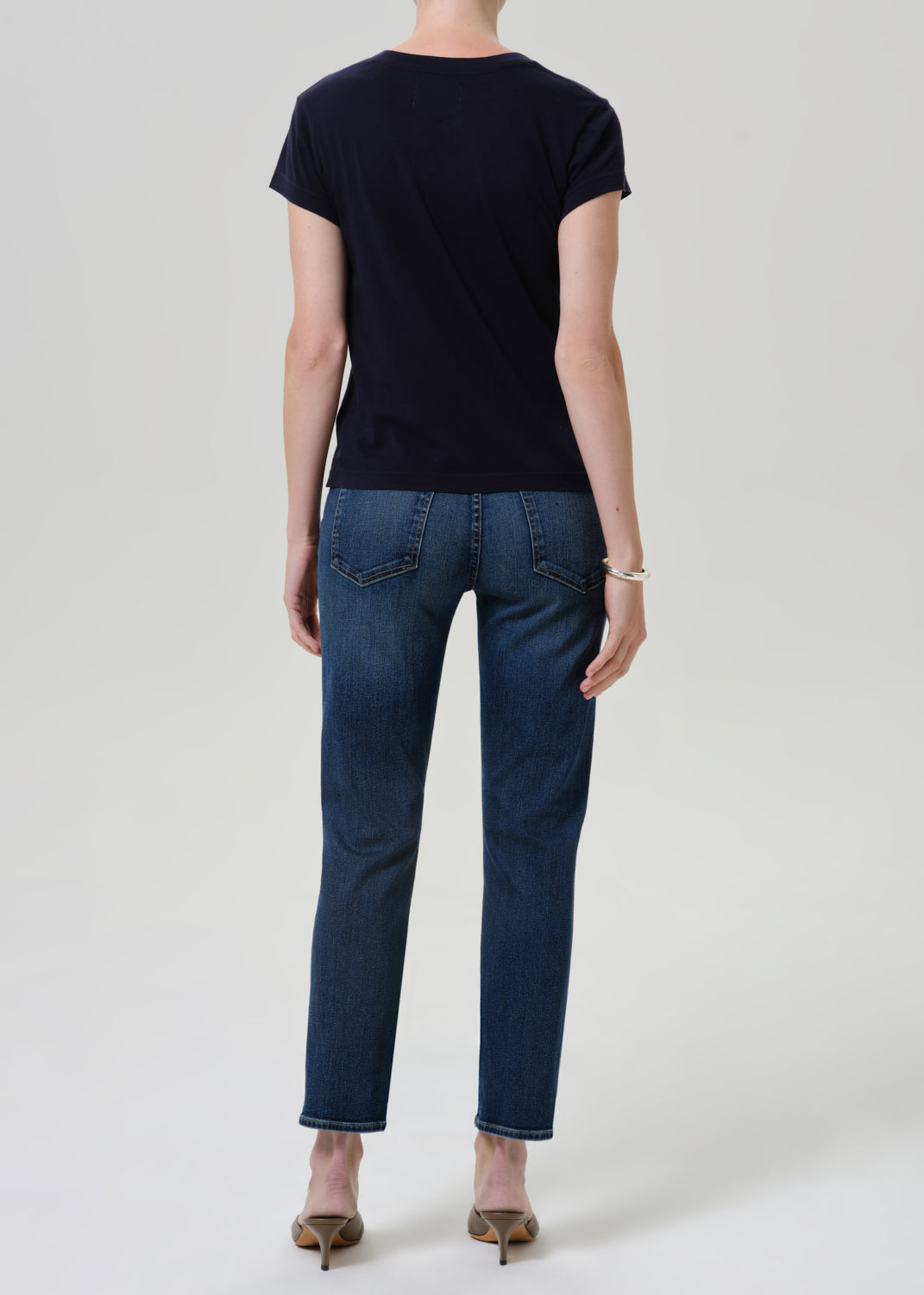 Person wearing dark blue straight leg cropped jeans with a navy short sleeve crewneck t-shirt and heels, back full body view on a grey background.