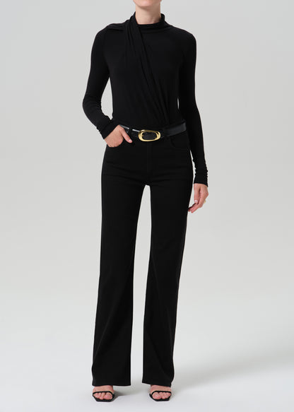 A model wearing a black long-sleeve draped mock-neck top and matching black wide-leg trousers, styled with a black belt featuring a gold oval buckle and black strappy heels, against a grey background.