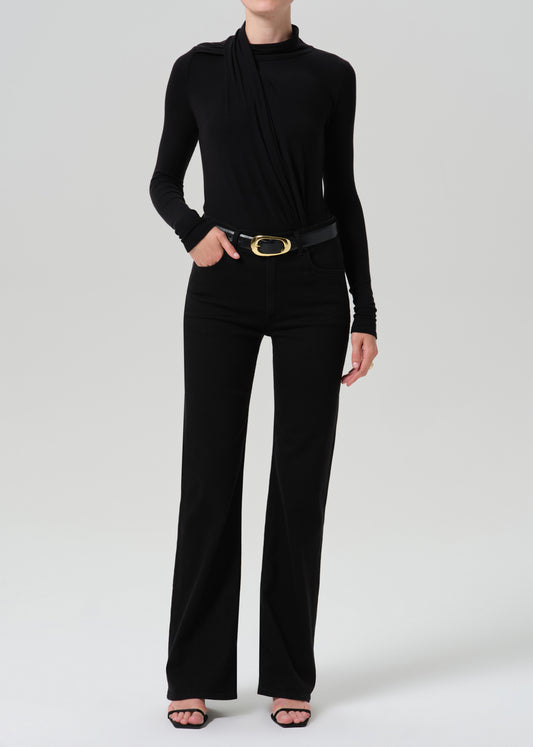 A model wearing a black long-sleeve draped mock-neck top and matching black wide-leg trousers, styled with a black belt featuring a gold oval buckle and black strappy heels, against a grey background.