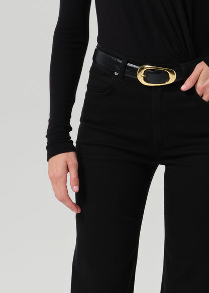 A close-up of a person wearing black high-waisted jeans and a black belt with a prominent gold oval buckle, paired with a black long-sleeve top against a grey background.