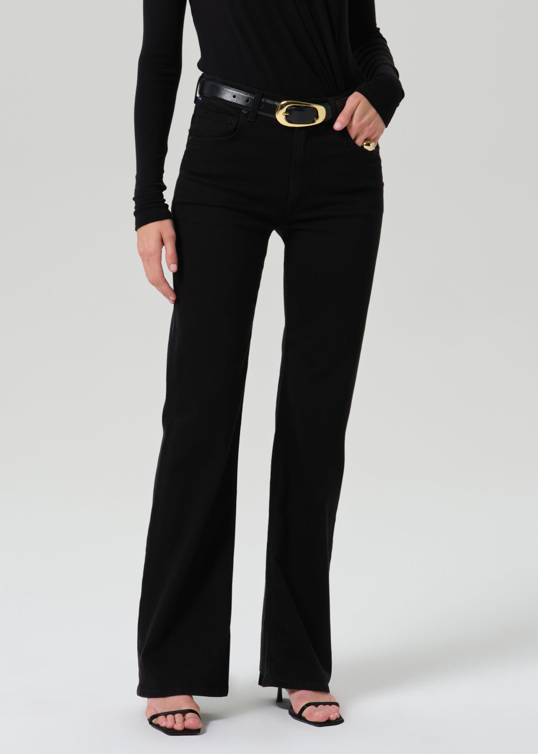 A model wearing black high-waisted flared jeans with a black leather belt featuring a gold oval buckle, paired with a black long-sleeve top and black sandals, against a grey background.