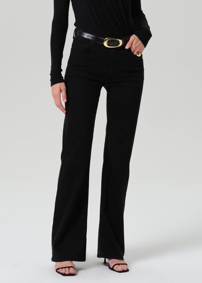 A model wearing black high-waisted flared jeans with a black leather belt featuring a gold oval buckle, paired with a black long-sleeve top and black sandals, against a grey background.