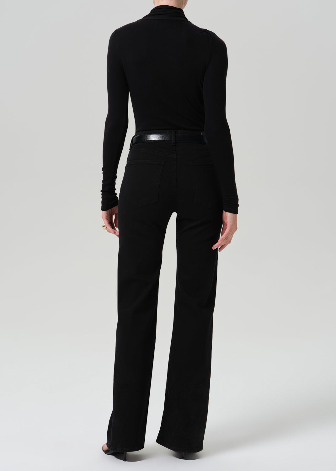 A model shown from the back wearing a black long-sleeve mock neck top and black wide-leg trousers, paired with a black belt, against a grey background.
