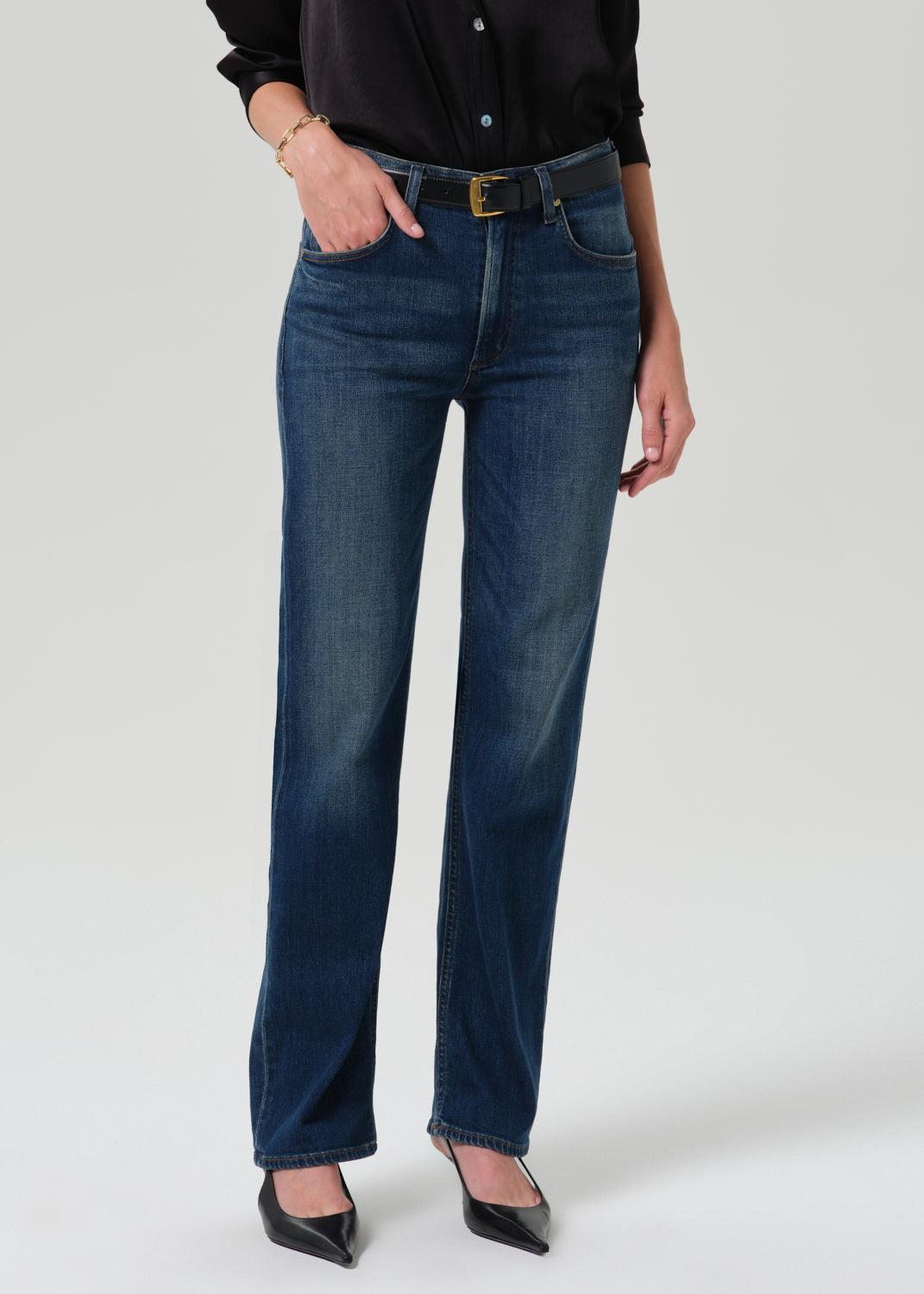 Person wearing a black long sleeve button front blouse with dark indigo straight leg jeans on a grey background.