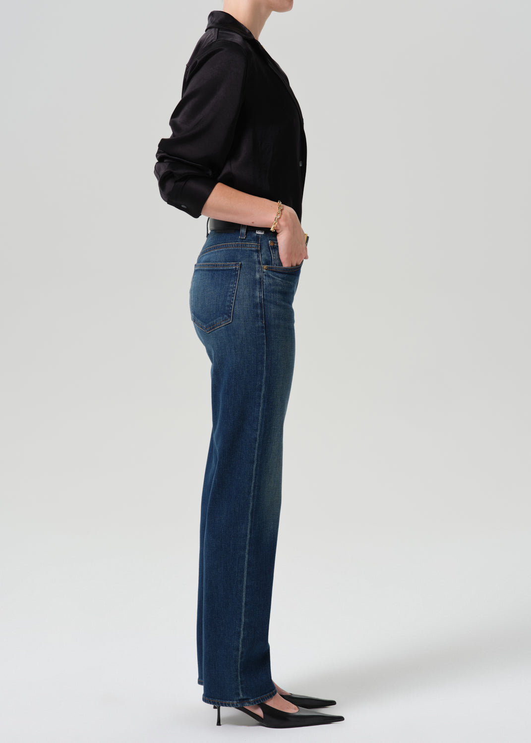 Person wearing a black long sleeve button front blouse with dark indigo straight leg jeans viewed from the side on a grey background.