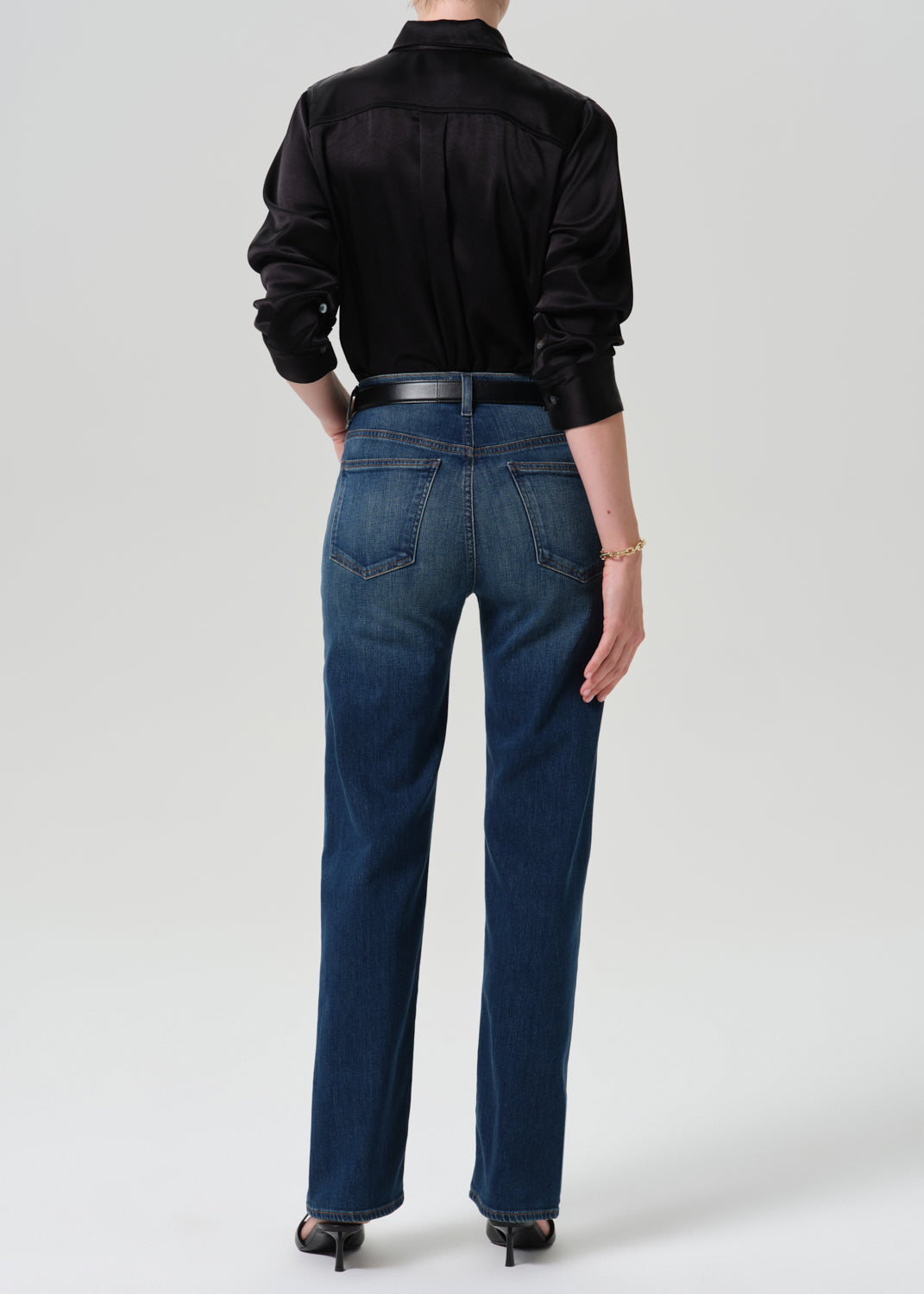 Person wearing a black long sleeve button front blouse with dark indigo straight leg jeans viewed from the back on a grey background.
