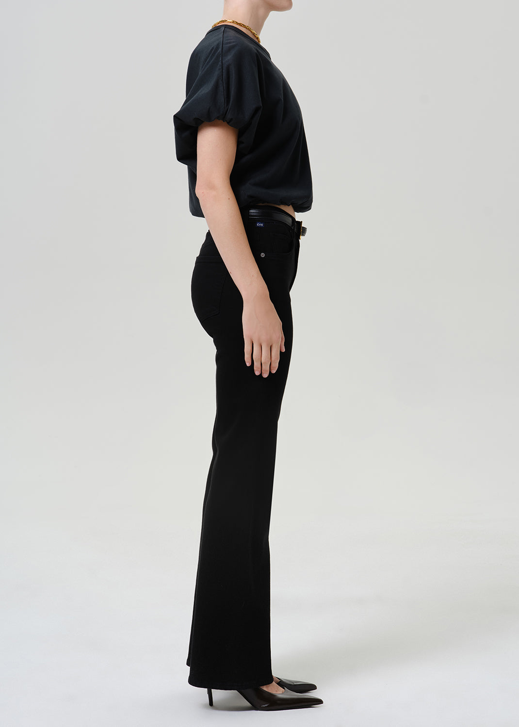 Woman wearing black straight leg pants with a black short sleeve top viewed from the side on a grey background.