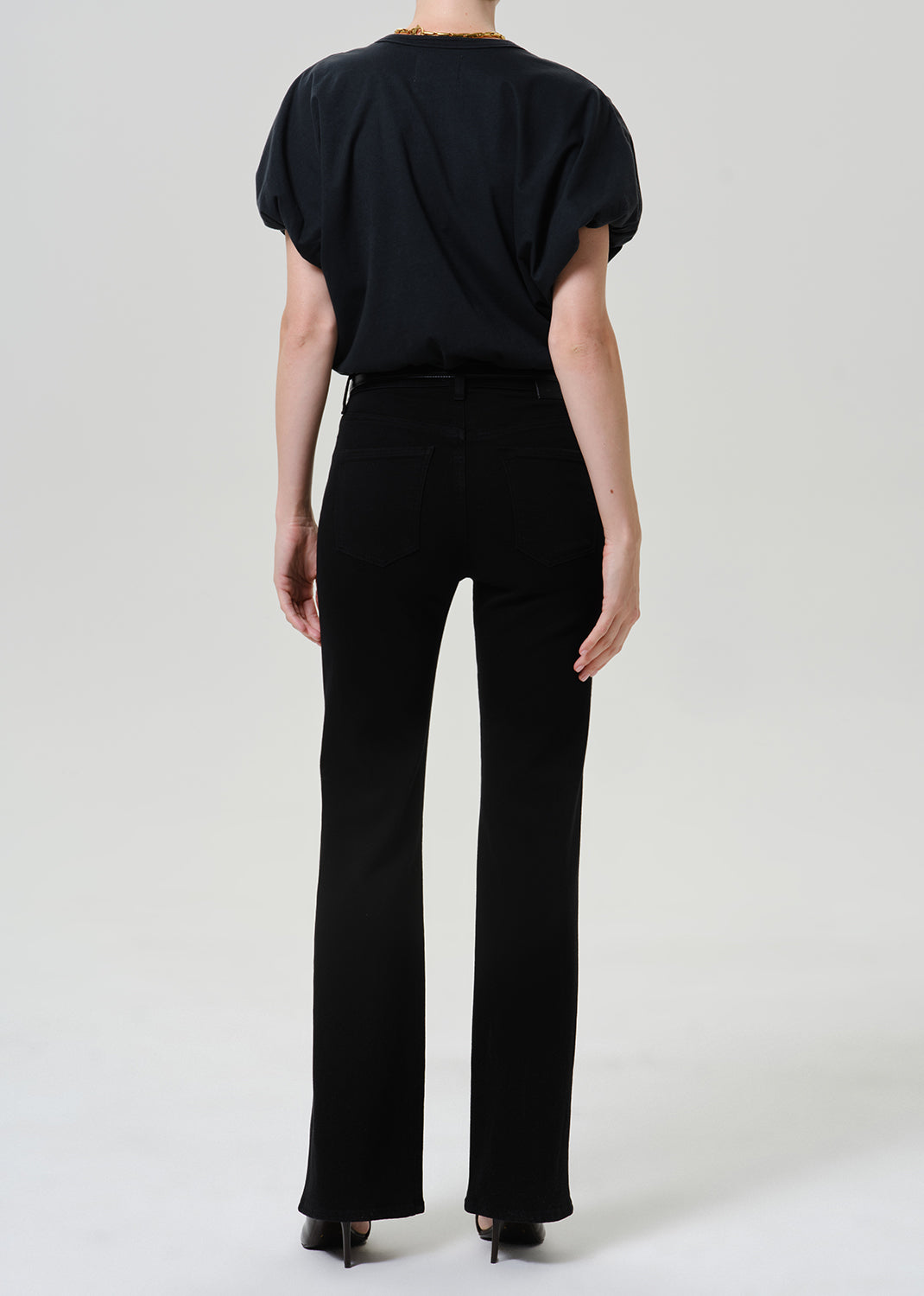 Woman wearing black straight leg pants with a black short sleeve top viewed from the back on a grey background.