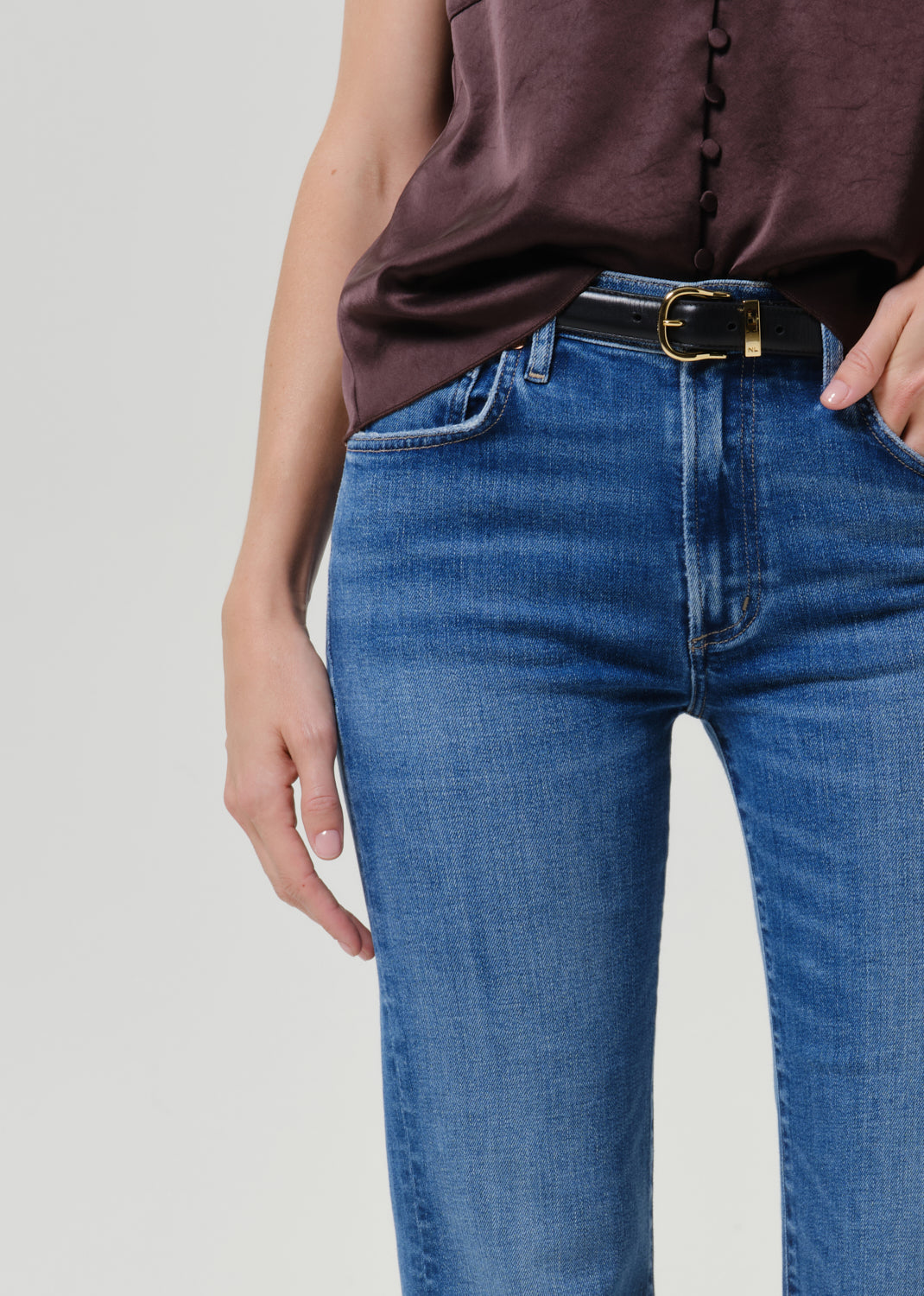 Close-up of a person wearing blue denim jeans with a black belt and gold buckle, paired with a dark brown top, against a light gray studio background.