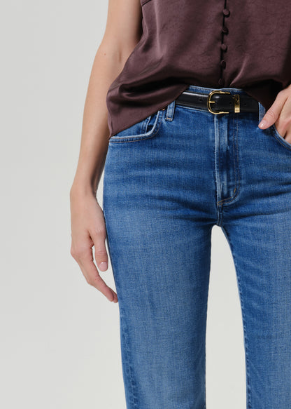 Close-up of a person wearing blue denim jeans with a black belt and gold buckle, paired with a dark brown top, against a light gray studio background.