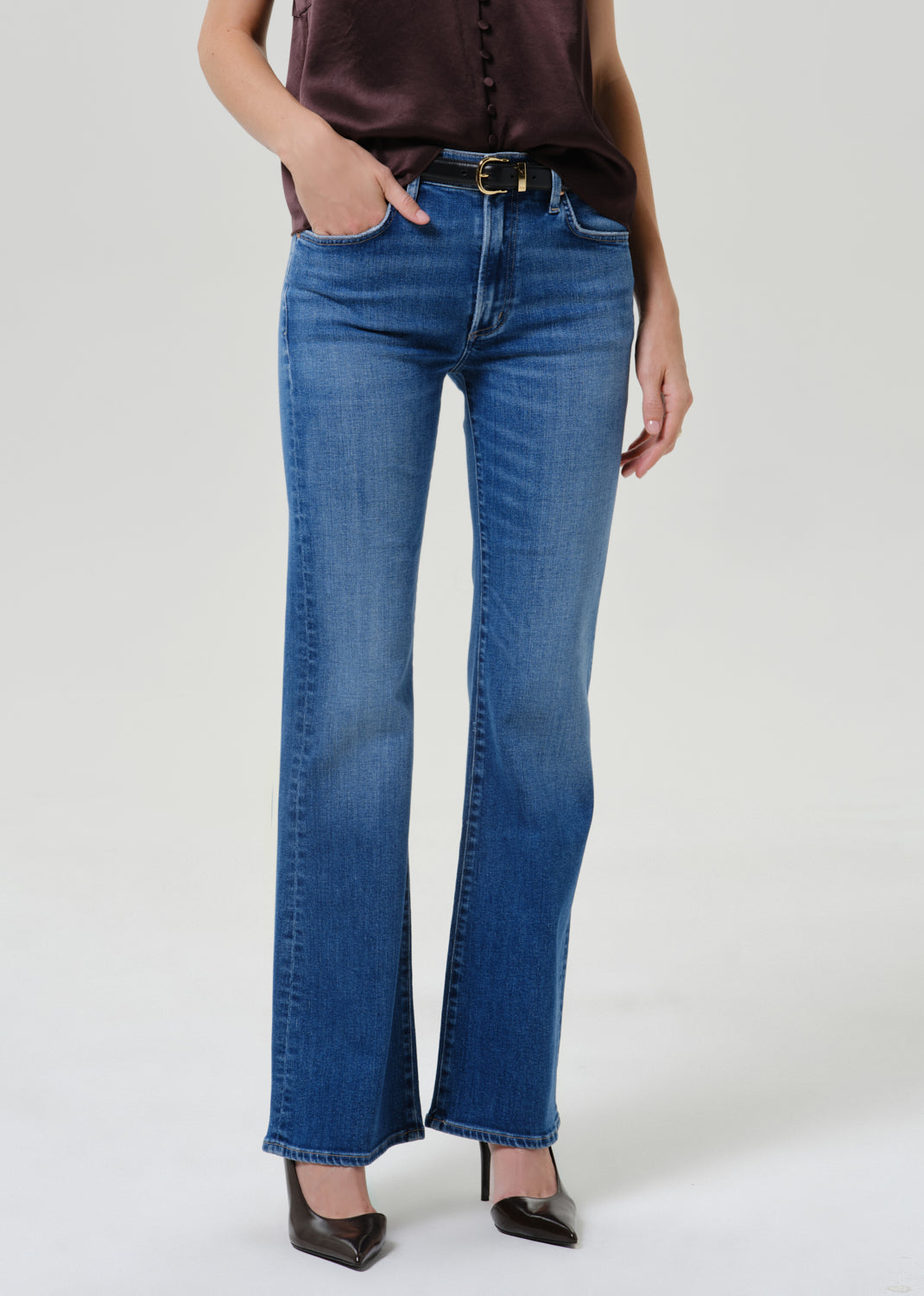 A model wearing a pair of medium-wash blue flare jeans with a dark brown satin top and black pointed-toe heels, standing against a light gray studio background.