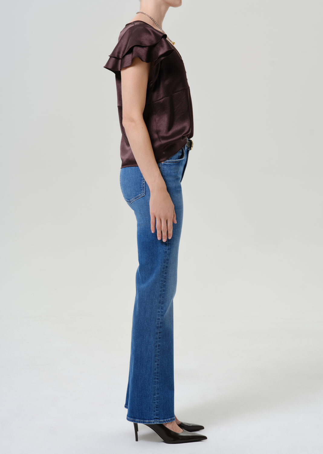 A model shown in profile wearing a dark brown ruffled short-sleeve top, blue flare jeans, and black pointed-toe heels against a light gray studio background.