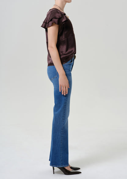 A model shown in profile wearing a dark brown ruffled short-sleeve top, blue flare jeans, and black pointed-toe heels against a light gray studio background.