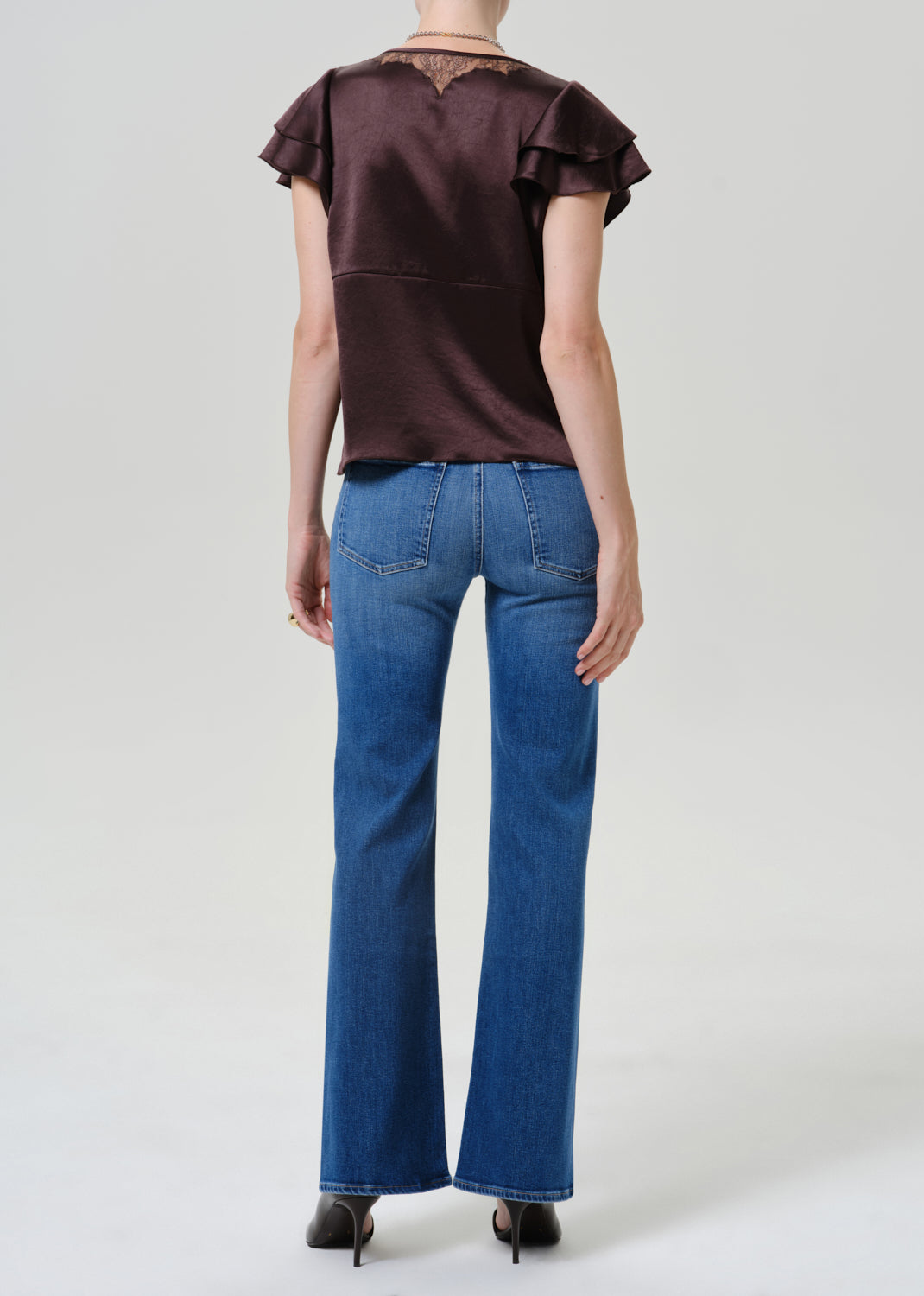 A model shown from the back wearing a dark brown ruffled short-sleeve top and blue flare jeans, standing against a light gray studio background.