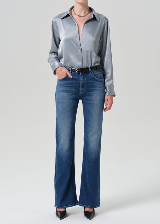A model wearing a metallic silver satin button-down shirt and blue wide-leg jeans, standing against a grey background.