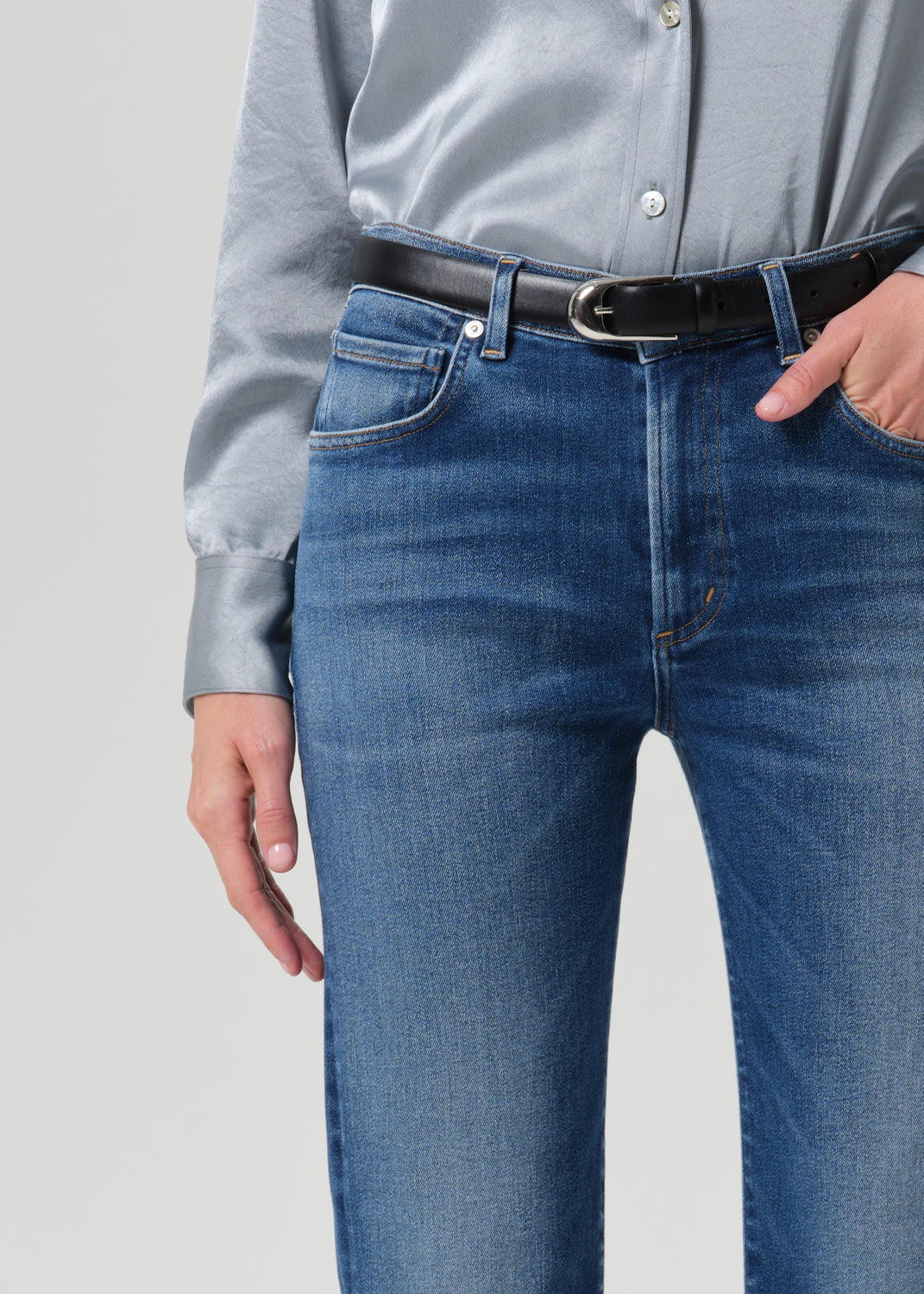 Close-up of a person wearing blue jeans with a black belt, a light blue satin shirt, and a silver belt buckle, against a grey background.