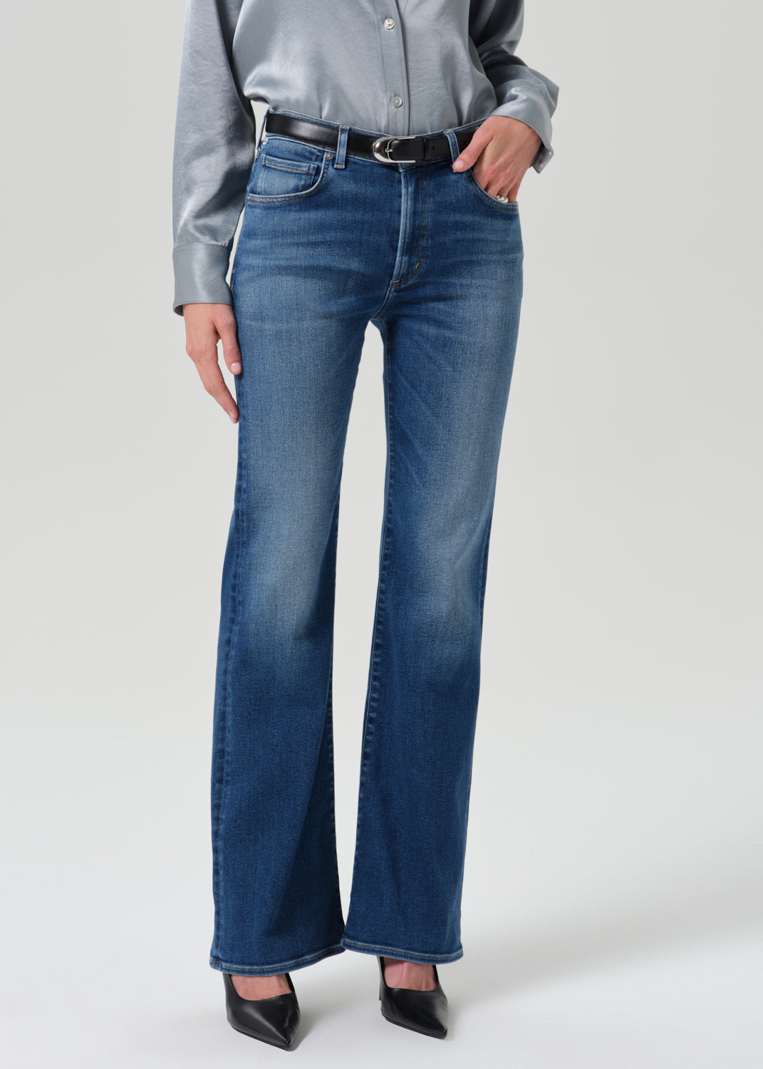 A model wearing a pair of high-waisted, wide-leg blue denim jeans with a black belt, paired with a gray satin shirt and black pointed-toe heels, photographed against a grey background.