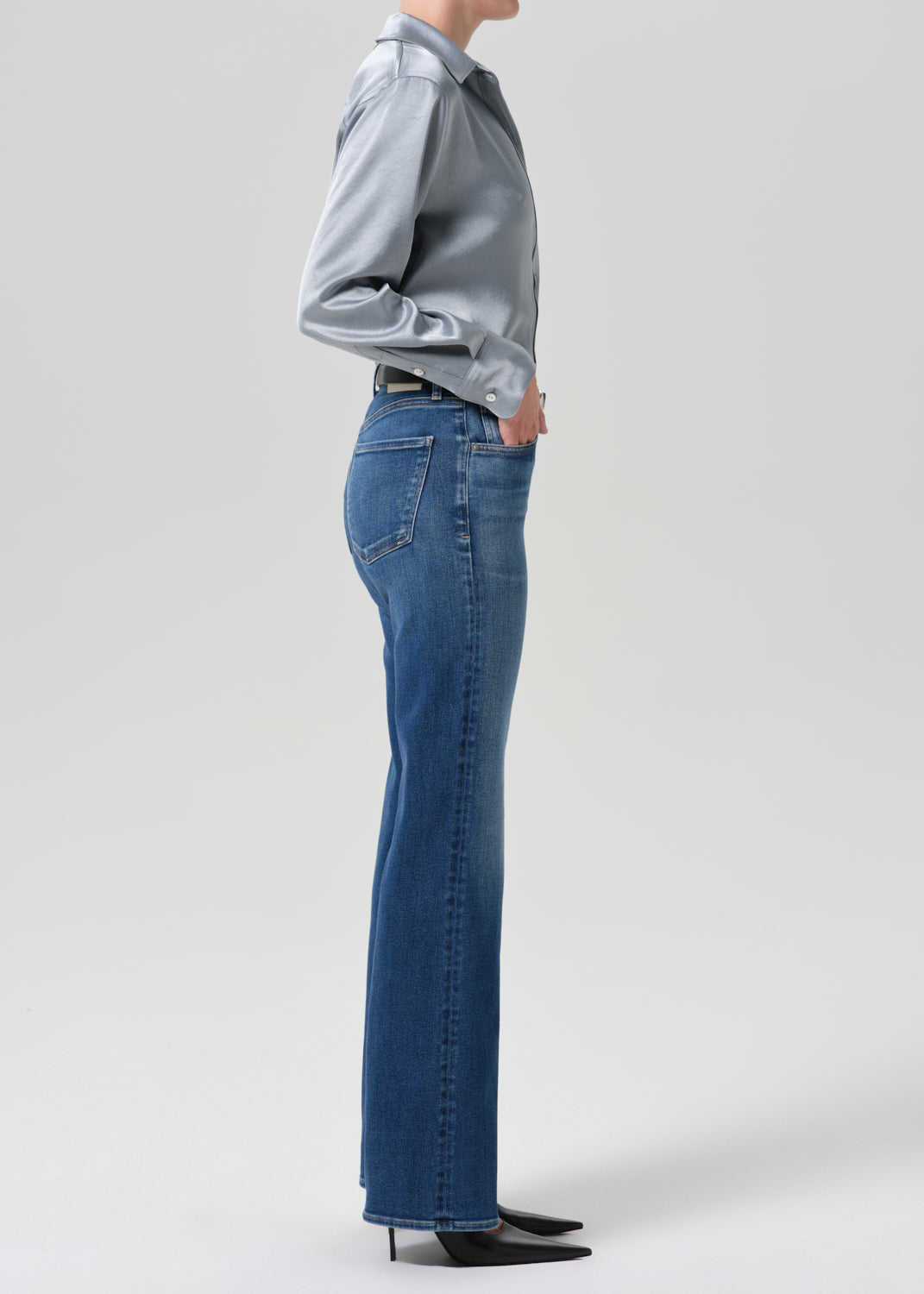 A side-profile studio shot of a model wearing blue high-waisted flare jeans, a light blue satin shirt, and black pointed-toe heels against a grey background.