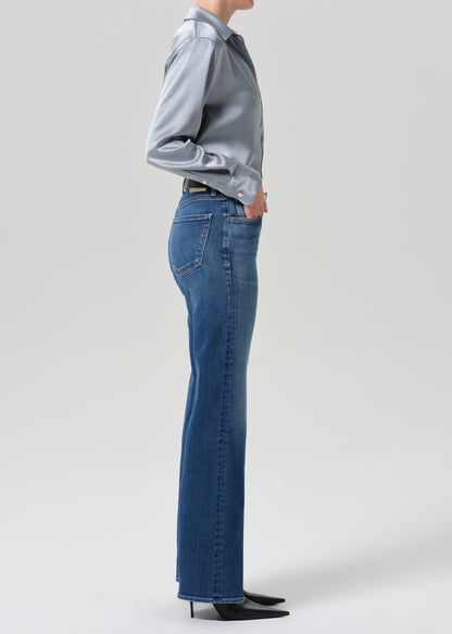A side-profile studio shot of a model wearing blue high-waisted flare jeans, a light blue satin shirt, and black pointed-toe heels against a grey background.