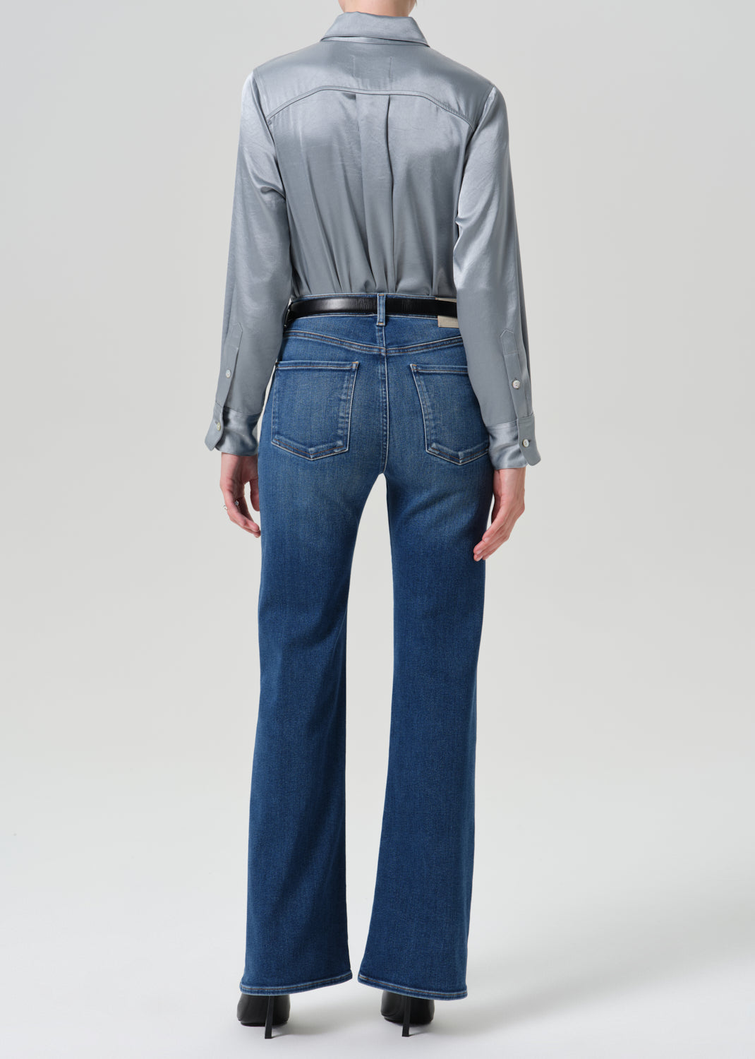 A model shown from the back wearing a shiny silver satin shirt tucked into blue flare jeans with a black belt, photographed against a grey background.
