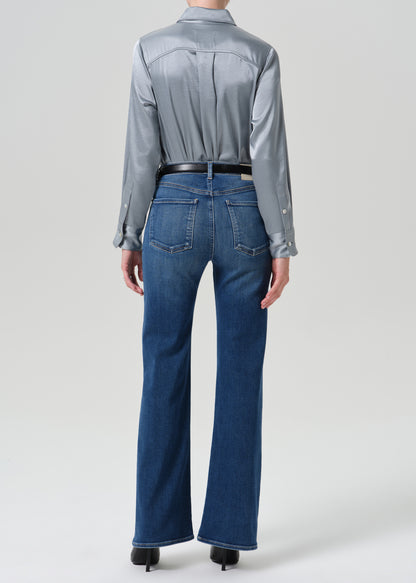 A model shown from the back wearing a shiny silver satin shirt tucked into blue flare jeans with a black belt, photographed against a grey background.