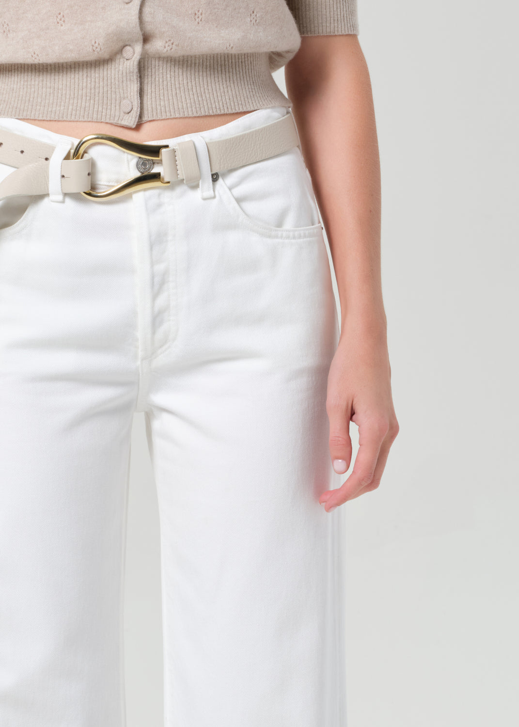 Person wearing a beige cardigan and white jeans on a plain background