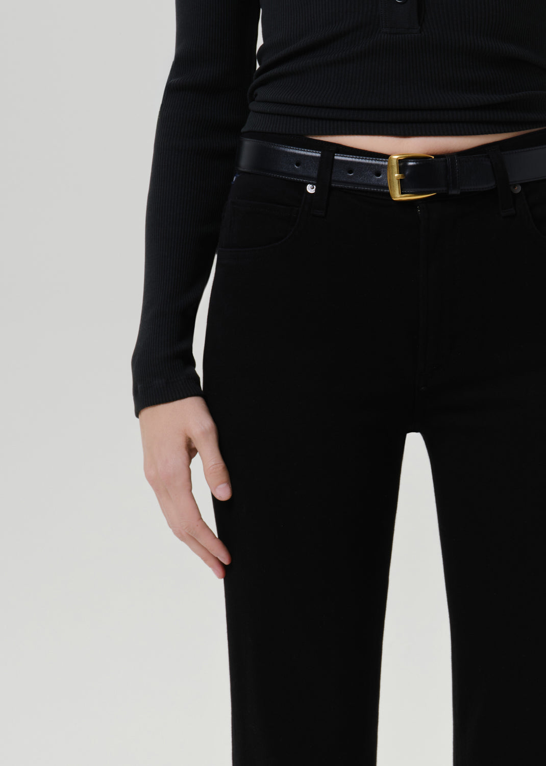 A close-up of a model wearing black high-waisted straight-leg jeans, a black leather belt with a gold buckle, and a black ribbed long-sleeve top against a grey background.