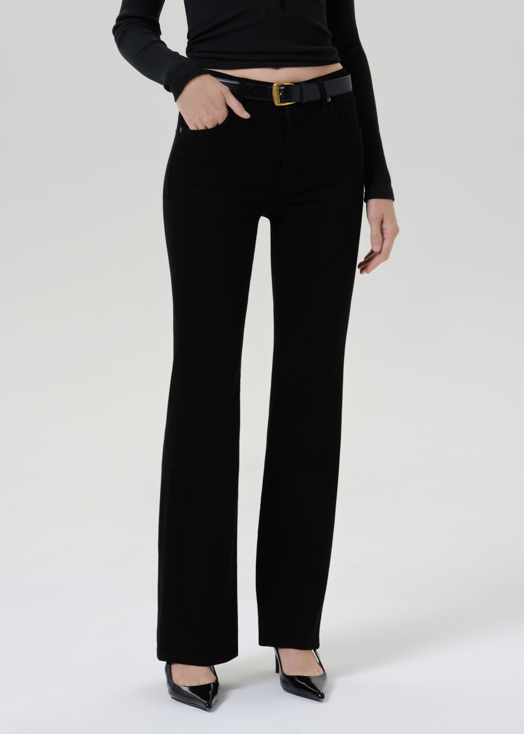A model wearing black wide-leg trousers with a black belt and black pointed-toe heels, shown from the waist down against a grey background.