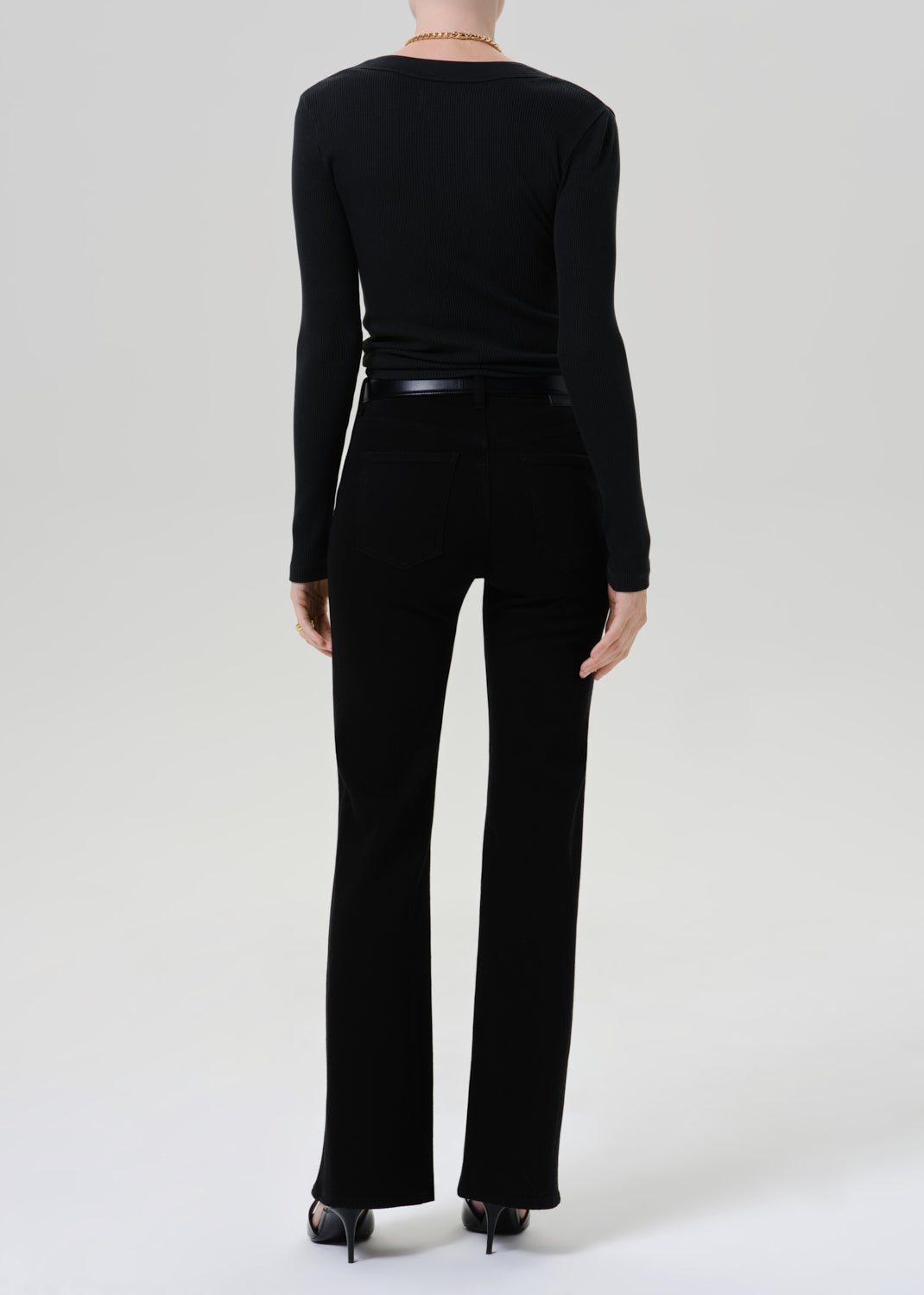 A model shown from the back wearing a black long-sleeve top and black flared pants, paired with black heels, against a grey background.