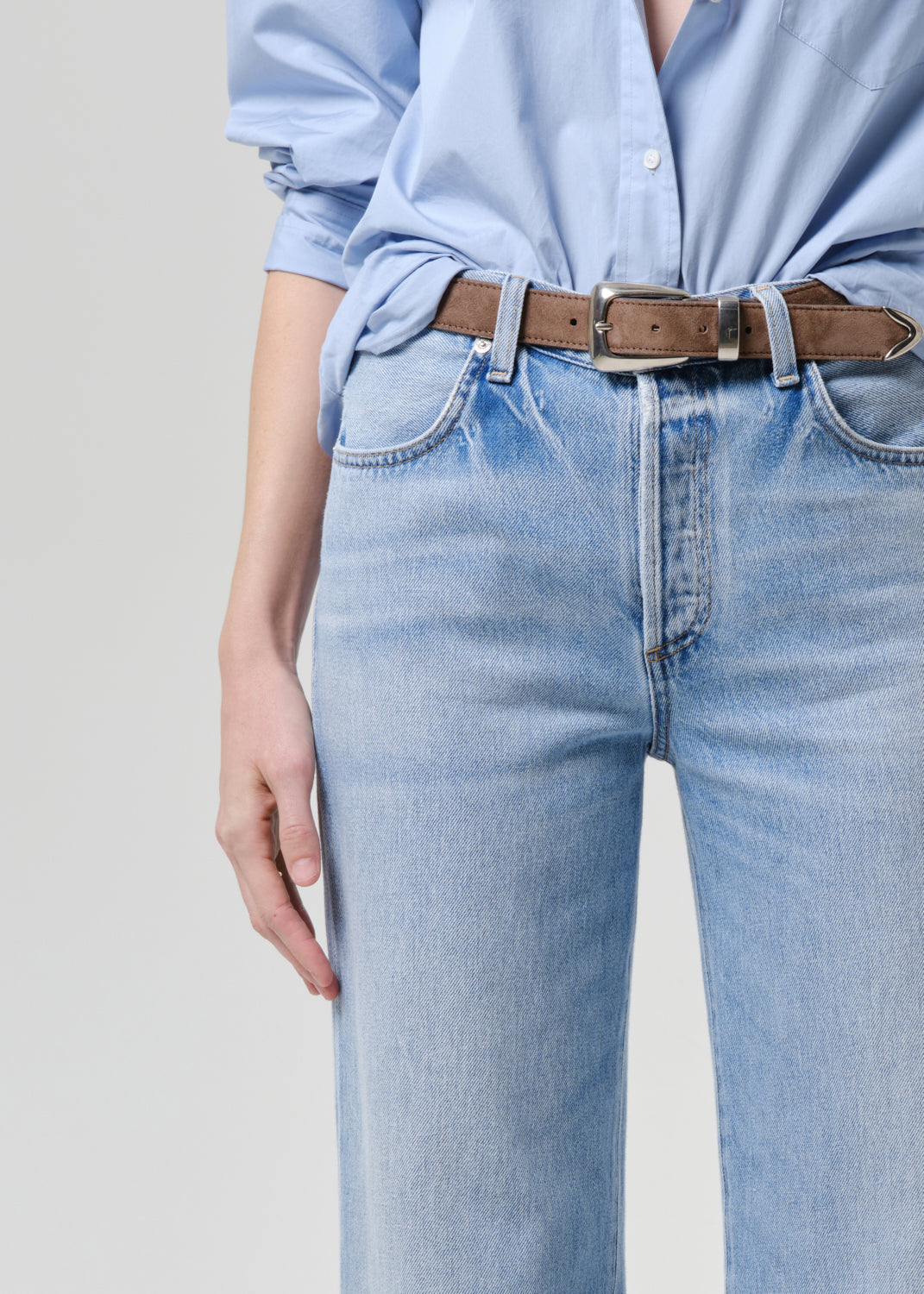 A close-up of a person wearing light blue jeans, a light blue button-up shirt, and a brown leather belt with a silver buckle, against a grey background.